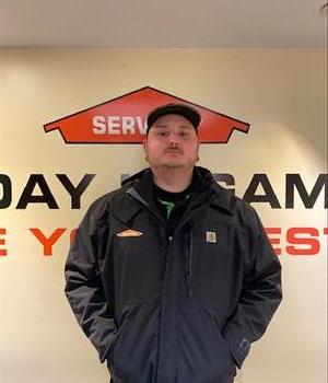 Younger male with a neatly trimmed mustache, wearing a ball cap and jacket bearing the SERVPRO emblem